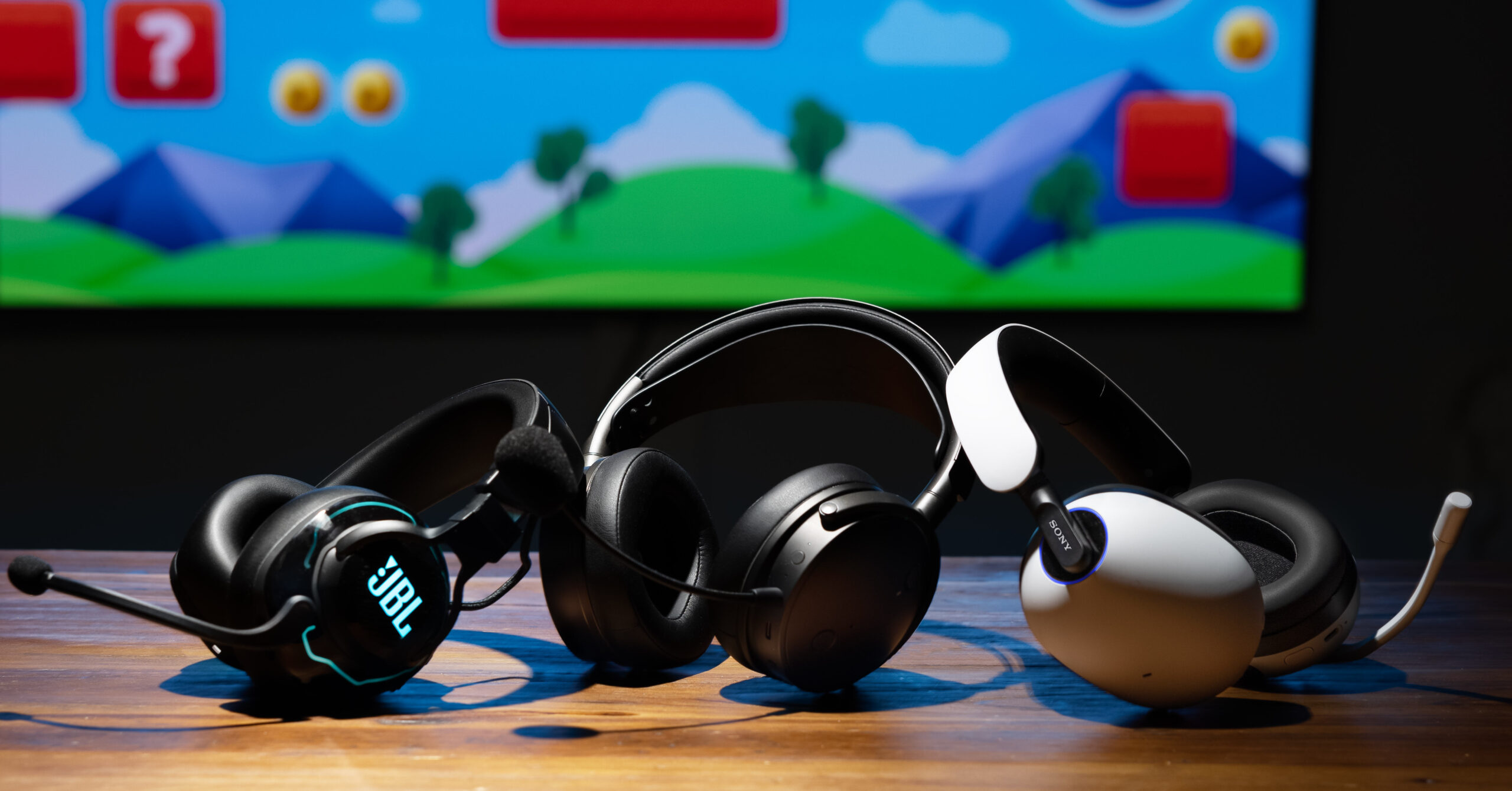 three gaming headsets arranged on a wooden table, with a colorful, cartoon-like background displayed on a screen behind them. The headsets include a JBL model on the left, a black wireless headset in the middle, and a white headset on the right