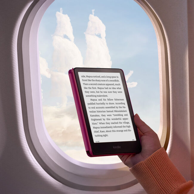 Person holding a Kindle Paperwhite e-reader in front of an airplane window, with fluffy clouds and a blue sky visible outside.